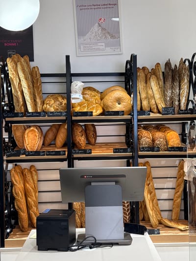 This image captures the interior of a boulangerie (bakery) in Asnières-sur-Seine, France, showcasing a wide selection of bread products. The scene is dominated by multiple black metal shelving units with light wooden shelves, displaying an array of freshly baked bread.
On these shelves, various types of bread are arranged: numerous long baguettes of varying crust colors (light to dark), round loaves, oval loaves, braided bread, and distinctive bagels. Many items are labeled with small black price tags in French, indicating types such as "Pain Italien," "Baguette Italienne," "Pain Turc," "Pain d'Orge," "Grand Boule," "Petit Boulot," "Pain Complet," "Pain Battu graines," "Pain de Seigle," "Pain Battu," "Pain de Maïs," and "Baguette des Prés."
In the foreground, a modern point-of-sale (POS) system is visible on a white counter. It consists of a grey monitor with a "DISH" logo, angled away from the viewer, and a black POS printer located to its left. Electrical cords are visible near the base of the monitor.
The walls are white. On the upper right wall, a framed poster features a large pile of white flour with a red "R" emblem and French text stating, "J'utilise une farine Label Rouge, garantie de qualité supérieure." A partially visible sign on the upper left wall reads "FÉROCEMENT BONNE!" A large, spherical white light fixture hangs from the ceiling in the upper left. No individuals are present in the image. The environment appears clean and well-lit.