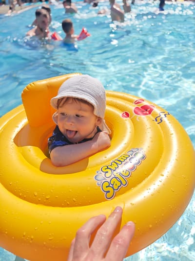 A baby wearing a white sun hat and a dark blue swimsuit is sitting in a bright yellow inflatable swim ring in a swimming pool. The baby is sticking their tongue out, with a playful expression. The swim ring has red crab graphics and the words "Swim Safe" printed on it. Water droplets are visible on the surface of the swim ring. A person's hand, likely an adult, is reaching out to touch the swim ring from the bottom right of the frame. In the background, other people are visible in the pool, some with arm floaties, suggesting a family outing or a public swimming area. The water is clear blue, and the sunlight appears bright, indicating it is likely daytime. The overall scene conveys a sense of summer fun and safety for the child in the water. The location is Georgsmarienhütte, Germany.