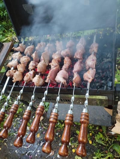 The image depicts a close-up view of a barbecue grill with numerous skewers of meat being cooked over glowing coals. Smoke rises from the grill, indicating active cooking. The skewers are made of metal with decorative wooden handles, arranged in several rows. The meat on the skewers appears to be cubed and is in various stages of cooking. The grill itself is a dark metal structure with a lid partially open. The background shows foliage and greenery, suggesting an outdoor setting, possibly in a rural or park-like area. The ground around the grill is covered with dirt, grass, and fallen leaves. The overall scene captures the preparation of a meal outdoors.