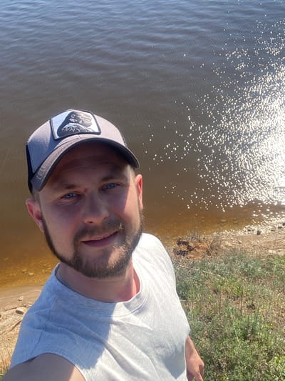The image shows a selfie taken outdoors, likely by a man with fair skin, blue eyes, and a beard, wearing a white t-shirt and a light gray baseball cap with a patch on the side. The background includes a body of water with reflective sunlight, and some vegetation along the shoreline. The photograph appears to have been taken in daylight with bright lighting conditions. Based on the details given, the location is Шуран, Russia.