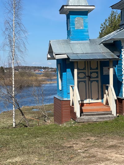 The image shows a blue wooden building next to a body of water. The building has a metal roof, decorative trim, and a door with white panels. A small sign is visible above the door. The water is likely a lake or wide river, with trees and houses visible on the opposite shore. The foreground shows grass, bare tree branches, and a birch tree. Based on the sign mentioning "Chapel P. 1905" (translated), it is likely in or near Чернышово, Russia.
