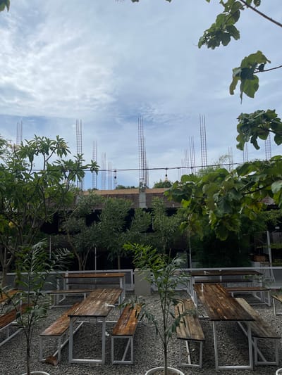 This image depicts an outdoor seating area with wooden tables and benches on a gravel ground. In the background, a building is under construction, with exposed rebar and brickwork visible. Lush green trees and foliage surround the seating area, with some branches extending into the frame from the top. The sky is cloudy with patches of blue. The scene appears to be set in a cafe or restaurant located in Pulokiton, Indonesia, given the tropical vegetation and the style of outdoor seating often found in such establishments. No people are visible in the image.