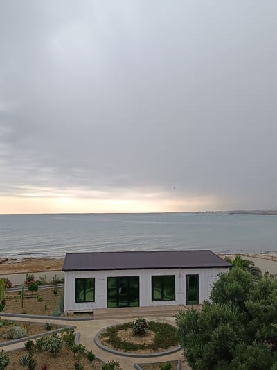 A modern, white, rectangular building with a dark, sloped roof is situated on a coastal property. The building features large, dark-tinted windows. In front of the building, there is a landscaped area with pathways, sand, and various plants, including shrubs and succulents. Beyond the landscaped area and building, a calm, blue sea stretches to the horizon. The sky above is overcast, with the sun faintly visible as a diffused light source near the horizon, casting a soft glow over the water. In the distance, along the coastline, there are structures that appear to be piers or possibly industrial buildings. The overall scene is one of a serene, coastal environment, possibly a resort or a private residence, located in the Sabunchu Raion of Azerbaijan.