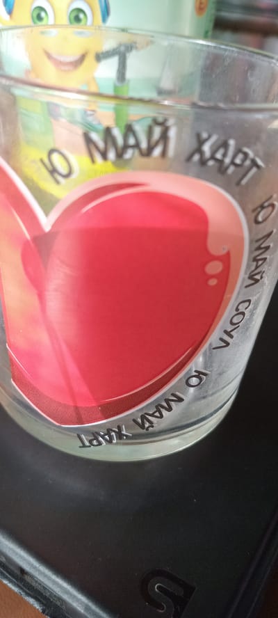 The image shows a clear glass with text and a heart design. The text "10 МАЙ ХАРТ" (10 May Hart) is printed on the glass in white lettering. Part of a yellow cartoon character with green eyes is visible in the background. The glass is resting on a dark, textured surface. The location in the city of Koviaky, Ukraine is not ascertainable from the image.