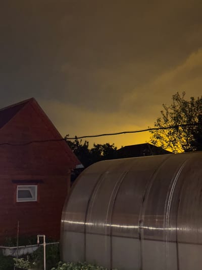 The image depicts an exterior nighttime scene in what appears to be the city of Mayak, Russia, given the context. The focus is on a red brick building with a single, small square window with the dark interior space visible through it. A large, shiny, cylindrical structure, possibly a metal storage tank or silo, dominates the right side of the frame. Overhead electrical wiring is visible against the background sky and near tree branches. The sky appears to be overcast and illuminated by an amber glow, suggesting artificial lighting. The ground is obscured by foliage.
