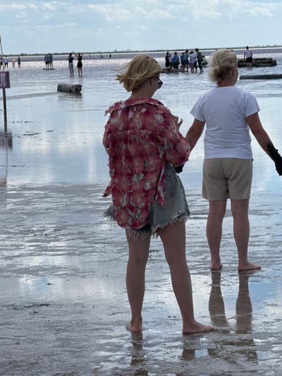 This is a medium shot of two women standing in shallow water at a salt lake in Bursol, Russia. The woman in the foreground is facing away from the camera, wearing a red and white patterned shirt and denim shorts. She has blonde hair and is barefoot. The woman in the background is also facing away from the camera, wearing a white t-shirt and beige shorts. She has blonde hair and is barefoot. Both women appear to be holding hands or reaching towards each other. The water is shallow and reflects the cloudy sky. In the background, other people can be seen standing in the water and on wooden platforms. The ground is covered in salt deposits.