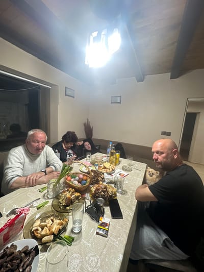 A group of people is gathered around a table laden with food, likely in a home setting. There are at least five individuals visible, ranging in age. A man with a shaved head and a black shirt is looking at the food on the table, where a small dog is also present. There is a table covered with a patterned tablecloth, holding plates of food, including meat dishes, bread, and a basket of Easter eggs. There are also beverages like water, some electronic devices like a phone, and other personal items on the table. The room has a somewhat rustic feel with exposed wooden beams and a bright light source. The location is Vinarovo, Bulgaria. The general atmosphere suggests a family gathering or meal.