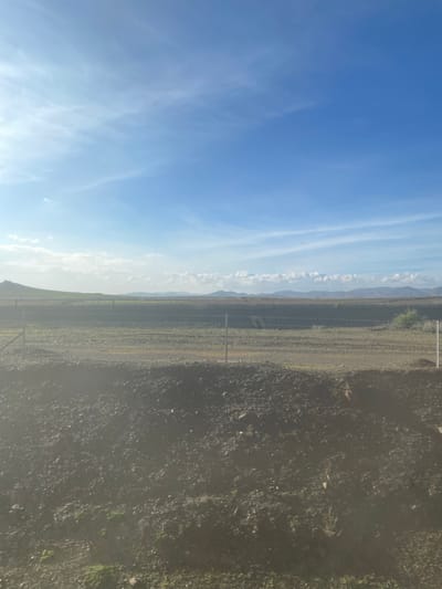 The image displays a vast, open landscape under a bright, clear sky. When viewed horizontally, the sky occupies the upper portion, a distant mountain range lies at the horizon, and various ground textures fill the midground and foreground.
The sky is a vivid blue, predominantly clear, with delicate, wispy white clouds stretching across it, particularly visible on the left side. These clouds are thin and elongated, suggesting calm atmospheric conditions.
Along the horizon, a long line of distant mountains or hills is visible, appearing somewhat hazy and light-toned, fading into the sky. This mountain range extends across the entire width of the image.
Below the mountains, the midground features a flat expanse of land. This area shows variations in color, from lighter greens to browns, suggesting either sparse vegetation or cultivated fields. A series of thin, vertical posts, likely part of a fence, runs horizontally across this section, indicating a boundary.
The foreground, particularly on the right side, is composed of darker, rougher terrain. This area appears to be uncultivated ground or a dirt track, with scattered stones and dry, dark patches of low-lying vegetation.
The scene depicts a bright, sunny day, likely during the morning or afternoon. The weather is clear with excellent visibility. No people or specific activities are present. The arid or semi-arid appearance of the landscape, with distant mountains and open plains, aligns with the context of Jbilate, Morocco. There is no visible text in the image. The overall impression is one of peacefulness and expansive natural scenery.