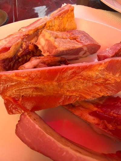 A close-up, overhead shot displays multiple cuts of raw, pinkish-red meat, likely pork, on a white plate. The meat varies in size and thickness, with some pieces showing layers of fat and muscle. The lighting casts a reddish hue on some portions of the meat, suggesting it might be under a heat lamp. The background is blurred, but hints of red and white suggest a market stall or deli counter. There are no people or visible actions depicted in the image. The scene is devoid of any context relating to the city of Amzara, Abkhazia.