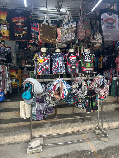 The image shows a street stall in Ban Thai–Changlon, Thailand, displaying a variety of clothing and bags. The stall features t-shirts with graphics related to motorcycling teams like Ducati and Kawasaki, as well as shirts with local themes such as "Hatyai Thailand" and "Songkhla." Several large bags with intricate elephant patterns are hung above the clothing. In the foreground, a metal rack holds folded textiles, including patterned fabrics and a stack of t-shirts and towels. The stall is set up on concrete steps leading to a storefront, with a wet ground surface suggesting recent rain. No people are visible in the image.