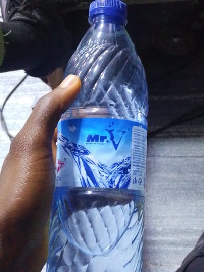 In Osogbo, Nigeria, a person's hand is holding a clear plastic bottle of "Mr.V Premium Water." The bottle has a blue cap and a label with blue and white graphics, including the brand name and images of water. The background is dark and indistinct, with some textured surfaces visible. The scene appears to be an indoor or shaded outdoor environment.