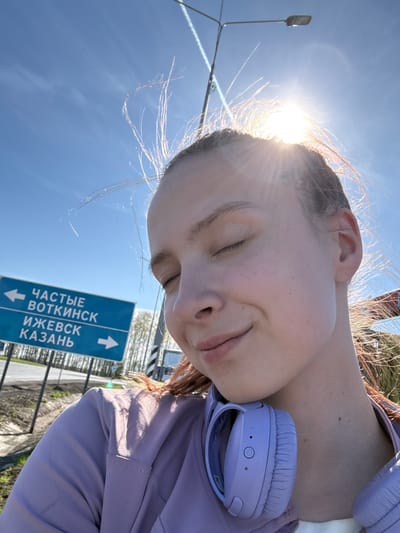 A young woman with fair skin and reddish hair is facing away from the viewer, head tilted to the side with eyes closed and lips slightly pursed. She is wearing a lavender-colored jacket and has lavender headphones around her neck. Behind her is a clear blue sky with the sun shining brightly, causing flares in the image. A light post is visible to the left, and a road sign is on the right. The sign is blue with white lettering in Russian, indicating directions to ЧАСТЫЕ (Chastye), ВОТКИНСК (Votkinsk), ИЖЕВСК (Izhevsk), and КАЗАНЬ (Kazan). The location is likely in the vicinity of Verkh-Sosnova, Russia.