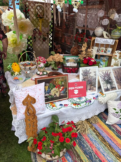 A stall at an outdoor market in Podlewkowie, Poland, is filled with handmade crafts and decorative items. The table is covered with a white lace-trimmed tablecloth, adorned with embroidered doilies, pillows, and framed artwork depicting fruits and lavender. Several baskets are present, one holding decorative Easter eggs and another containing various handcrafted items like a yellow chick and a grey knitted dog. A wooden carving of a tree-like figure stands upright on the table, and a pot of red flowers adds a vibrant splash of color. Behind the table, a backdrop of woven materials, lace curtains, and hanging decorative balls creates a rustic atmosphere. The ground is covered by a colorful striped rug, and wheat stalks are placed near the edge of the table. The scene suggests a festive or craft-themed event.