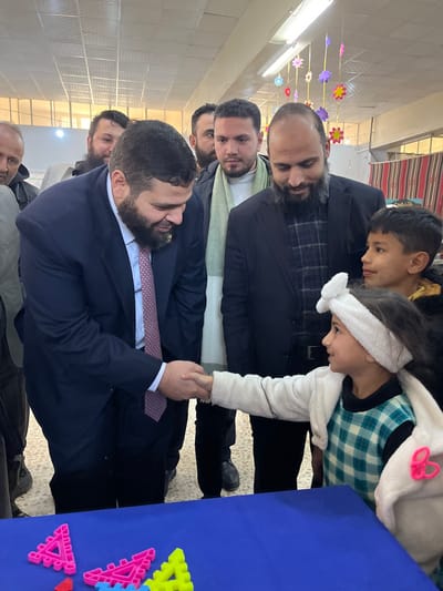 The image depicts an indoor scene in Kafr Batna, Syria, featuring seven individuals. A bearded adult male, wearing a dark suit, white shirt, and patterned tie, is shaking hands with a young girl. The girl, dressed in a light-colored top, plaid dress, and a white headband with a bow, is smiling and looking at the man. To the girl's right, a young boy in a dark jacket observes the handshake. Behind the girl, a bearded adult male in a dark jacket and plaid shirt also observes the interaction. Three other adult males are visible in the background: one partially visible to the far left, another bearded individual centrally behind the handshake, and a third male to the right of the central figure wearing a light green scarf and a dark jacket. In the immediate foreground, a blue table displays several brightly colored plastic geometric shapes (pink, blue, yellow triangles). The environment consists of white walls, fluorescent ceiling lights, and colorful paper flower decorations suspended from the ceiling. A striped fabric is visible on the far right background.