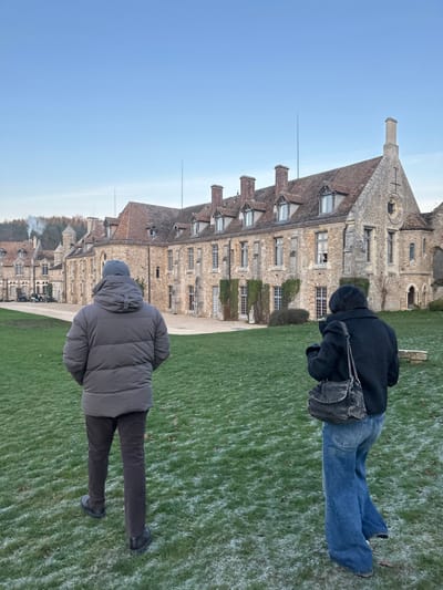 Two people stand on a grassy lawn in front of a large, historic stone building. The building is several stories high with many windows and a pitched roof. One person wears a dark jacket and blue jeans, while the other is dressed in a gray puffer coat and dark pants. The grass appears slightly frosty or damp. The sky is a clear blue.