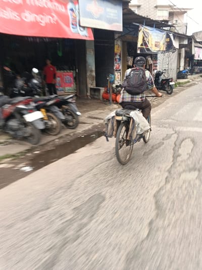 An overhead-angle, slightly blurred street-level photograph depicts a male individual riding a bicycle down a paved road in Dayeuhkolot, Indonesia. The rider, seen from behind, wears a plaid shirt, dark trousers, and a black baseball cap worn backward, along with a black backpack. The bicycle is dark-colored, equipped with a rear rack laden with light-colored, bundled items, and additional large packages or bags strapped to the sides of the rear wheel. The road surface is asphalt, exhibiting visible cracks and patches, with motion blur indicating movement.
To the immediate left of the road, a narrow paved sidewalk runs alongside a row of single-story commercial buildings. The closest building features a large red banner displaying partial Indonesian text including "matimu" and "alis dingin?", alongside graphics of "Oli Mak" branded oil bottles. Inside this establishment, multiple motorcycles are parked, and a person in a red shirt is visible standing. Adjacent to this, another shop displays a blue sign and a yellow banner with "WELDON" text. Outside this second shop, an orange air compressor, a small generator, and blue coiled hoses are visible on the sidewalk, suggesting a vehicle repair or tire shop. Several other motorcycles are parked along the sidewalk in front of these businesses. Further down the street, on both sides, more urban buildings are visible under an overcast sky.