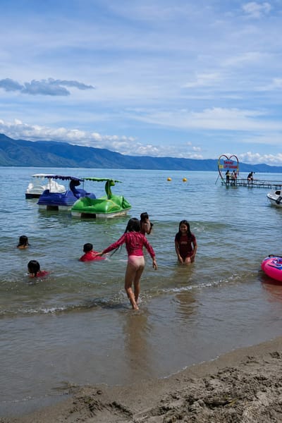 A group of children are swimming and playing in the shallow waters of a lake, near the shore. In the foreground, two girls in pink and maroon swimsuits are in the water, with one girl in the pink suit facing away from the camera, walking towards the deeper water. Several other children are visible in the water, some with their heads above the surface, also wearing swimsuits.

In the mid-ground, several colorful paddle boats are moored in the lake. Two are white and purple, and one is a bright green, shaped like a dinosaur. Yellow buoys are scattered in the water.

In the background, a wooden pier extends into the lake, with a large heart-shaped sign hanging above it. The sign has writing in Indonesian that appears to say "DANAU" and "ULUT TAWAR". Several people are standing on the pier, and a small white boat is docked to the right. Mountains line the horizon beyond the lake, under a bright blue sky with scattered clouds. The shoreline in the immediate foreground is a sandy beach with some debris. The overall scene depicts a recreational day at a lake in what appears to be a tropical or subtropical climate.