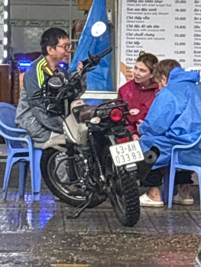 An outdoor scene in Ngũ Hành Sơn Ward, Đà Nẵng, Vietnam, depicts three individuals on a wet, tiled sidewalk. A grey and black motorcycle is centrally parked, displaying Vietnamese license plate '43-AH 033.83' on its rear fender. To the left of the motorcycle, an adult male, appearing of East Asian descent, wearing glasses and a grey tracksuit with yellow shoulder stripes, is seated on a blue plastic chair, looking towards the right. To the right of the motorcycle, a Caucasian female with light brown hair, wearing a maroon rain poncho, sits on a blue plastic chair, facing another individual in a blue rain suit, also seated on a blue plastic chair. All three appear engaged in conversation. A menu board with Vietnamese text and prices is visible in the background, behind the individuals on the right. The ground exhibits visible wetness and reflections, indicating recent precipitation. A blue flag or banner is partially visible in the upper background, behind the left individual.