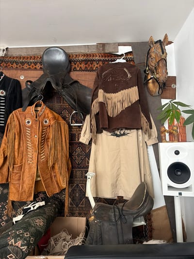 The image depicts a cluttered indoor space, possibly a storage area or workshop, with various equestrian and western-themed items. Prominently displayed are two saddles, one black leather saddle is hung high on the wall, and another darker brown leather saddle rests on a shelf lower down. Several garments are also visible, including a brown suede jacket with fringe and Native American-inspired embellishments, a tan-colored dress or skirt, and a black jacket with beaded details. A decorative wooden horse head with a bridle is mounted on the wall. Various rugs with geometric patterns are used as backdrops and to cover objects. A white speaker on a stand is positioned in the right side of the frame, next to a potted plant and a rustic vase. Boxes and rolled-up fabric are also present, contributing to the sense of disarray. The lighting suggests an indoor environment with artificial light. The scene is consistent with a collector's display or a storage space for vintage clothing and equestrian gear. There is no indication of the location in the city of Haarlem, Netherlands, or any people.