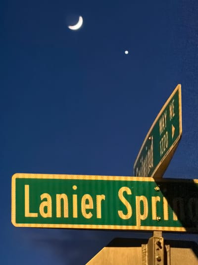 This is a low-angle, eye-level shot of two street signs at night in Buford, Georgia, United States. The primary sign, in the foreground, is green with yellow lettering that reads "Lanier Spr". It is bordered by a yellow reflective stripe. A second, curved green sign, in the background and to the right, has yellow lettering and appears to read "Southwind Way NE 1220". Both signs are mounted on a pole that is partially visible. In the dark blue night sky above, a thin crescent moon is visible on the left, and a small, bright white point of light, likely a planet, is visible to the right of the moon. The sky has a textured appearance, suggesting it may be a photograph with a canvas or similar texture applied.
