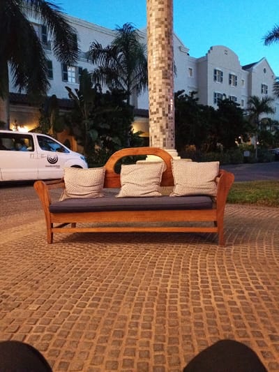 The image shows a wooden couch with three cushions positioned on a cobblestone patio. It is located in front of a building, likely a hotel, identified by a sign saying "Southern Sun" on a white van parked in the background. The building is multi-level and has large windows with white frames. Tropical greenery surrounds the building and there are trees in the background. The couch is positioned under the shade of a large palm tree, and there is a square-tiled column behind the couch. The sun is setting and the sky is a deep blue. The location, based on the provided coordinates, is likely the Southern Sun Maputo Hotel in Maputo, Mozambique.  No people are visible in the image.