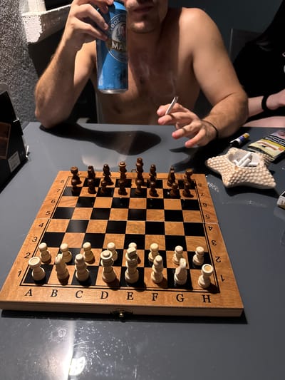 In Nea Potidea, Greece, a close-up shot focuses on a wooden chessboard set on a dark grey table. The chess pieces are arranged in their starting positions, with white pieces on ranks 1 and 2, and black pieces on ranks 7 and 8. The board is marked with letters A-H along the bottom and numbers 1-8 up the side. To the right of the chessboard is a star-shaped ashtray filled with cigarette butts. A man, partially visible from the shoulders up, is holding a can of beer in his left hand and a lit cigarette in his right. He is shirtless, and the environment appears to be indoors, possibly a living space or balcony, given the textured wall in the background. The lighting is dim, casting shadows on the table. Another person, wearing a black top, is partially visible on the right side of the frame, with their arm resting on the table.