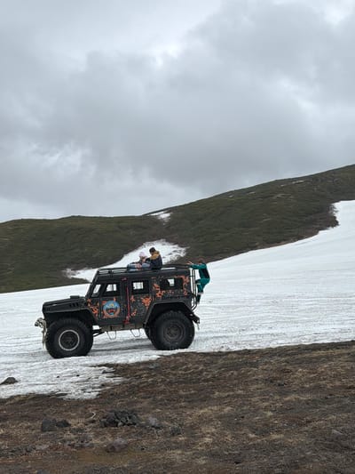 The image shows an all-terrain vehicle parked on a snowy incline with a barren, rocky foreground. Two people are seated on the roof of the vehicle, and another person is climbing onto the roof from the side. The vehicle is black with orange and white graphic designs. A green and blue tarp is draped over the side of the vehicle. The background features a green, grassy hill with patches of snow. The sky is overcast and cloudy. The scene appears to be a remote, outdoor location, likely in a mountainous region. The image does not provide specific details about the city of Petropavlovsk-Kamchatskiy, Russia.