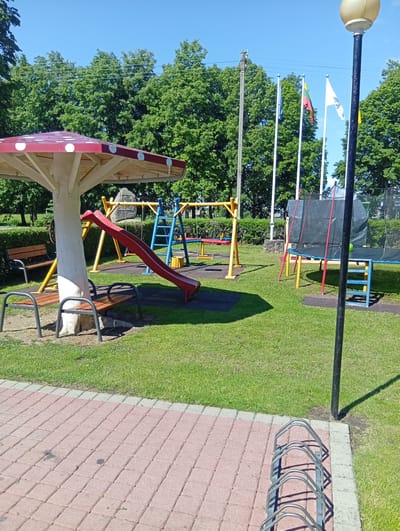 The image shows a children's playground on a sunny day in Dirvonėnai, Lithuania. The playground features a red mushroom-shaped canopy with white polka dots, a red slide, a swing set, and a trampoline with a black net enclosure. Benches are positioned near the mushroom canopy. In the background, several flags are visible on flagpoles, and a street lamp stands to the right of the image. The playground is surrounded by green grass and trees, with a paved area in the foreground that includes a bicycle rack. The sky is clear and blue.