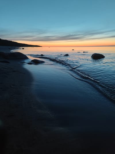A vertical, eye-level, medium shot captures a serene beach scene at dusk, with the camera positioned low to the ground, looking towards the sea. The foreground is dominated by dark, wet sand, reflecting the dim twilight sky. A gentle wave is captured in motion, lapping at the shore, its foam creating delicate patterns. Scattered along the beach and partially submerged in the water are several dark, rounded rocks, some larger than others. In the distance, a tree line of dark green foliage forms a silhouette against the horizon. The sky above is a gradient of colors, transitioning from a deep, clear blue at the top to soft hues of orange and pink near the horizon, indicating the setting sun. The overall mood is peaceful and tranquil.
