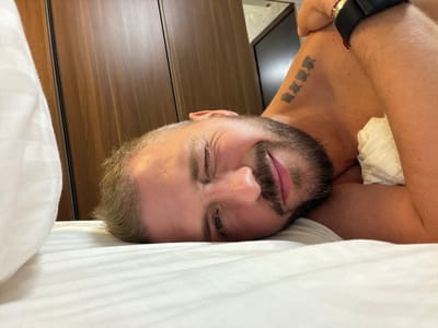 A man is depicted lying on his side on a white bed, looking directly at the camera with a playful and friendly expression. He is winking with his left eye and smiling broadly, conveying comfort and good humor. The man has a well-maintained dark beard and short, light-colored hair. He appears shirtless, with his bare left arm and shoulder visible. On his left upper arm, there is a tattoo composed of three distinct characters in dark Asian script. A dark-colored watch with a black band and a subtle red detail is worn on his left wrist.

The bed is neatly made with plain white sheets, and a white pillow is partially visible on the left side of the frame. In the background, a large dark brown wooden surface, possibly a wardrobe or paneling, dominates the space, indicating an indoor setting. Further back, hints of a lighter wall and another dark structure can be seen, consistent with a modern hotel room or apartment. The overall lighting is bright and even, illuminating the scene clearly. This relaxed and personal moment is set in what is understood to be Business Bay, United Arab Emirates.