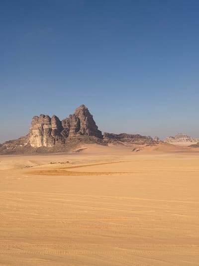 The scene depicts a vast desert landscape in Ash Shākirīyah, Jordan, under a clear, bright blue sky. The foreground and midground consist of expansive, flat, reddish-yellow sandy terrain. Multiple parallel tire tracks are visible across the sand, extending towards the background. A distinct, wide, curved streak of darker, disturbed sand traverses the midground from left to right. In the central background, a prominent, jagged massif of weathered rock formations rises, characterized by dark brown and grey strata and a notable lighter, sheer cliff face on its western portion. At the base of this main rock formation, in the mid-distance, a small cluster of indistinct structures, possibly tents, is discernible, accompanied by several small, dark, unidentifiable shapes. Further to the east in the distant background, smaller rock outcrops are visible. No explicit human figures or direct actions are clearly identifiable.
