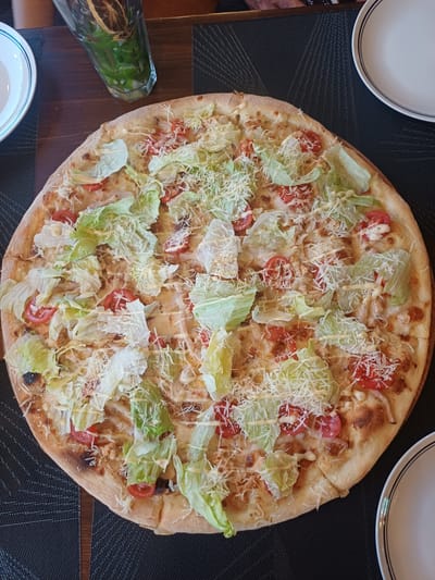 The image shows a close-up, top-down view of a whole Caesar salad pizza. The pizza is round with a thick crust, topped with shredded lettuce, cherry tomatoes, grated Parmesan cheese, and a drizzle of creamy dressing. The crust has some browned, slightly charred spots, indicating it's been baked. The pizza is placed on a dark, textured placemat, which is on a wooden table.
To the left of the pizza, there's a glass containing a beverage with visible green herbs and a slice of dried citrus fruit. In the upper left and bottom center of the image, parts of white plates with dark rims are visible, suggesting a dining setting. The lighting appears to be from overhead, casting soft shadows. The scene is likely indoors, possibly in a restaurant or home. There are no people visible in the frame, and no text is present. The overall impression is a prepared meal ready to be served or eaten, with a focus on the visually appealing presentation of the pizza.