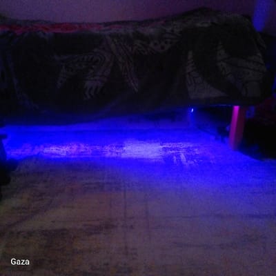 The image depicts an indoor environment in Kamera ward, Kenya, characterized by low light conditions and predominant blue illumination. A large, elevated object, consistent with a bed or similar furniture, occupies the upper half of the frame. This object is draped with a dark blanket featuring lighter, possibly tribal or abstract, patterns and a visible red border along its top edge. Intense blue light emanates from beneath this elevated object, casting a distinct blue glow across the floor surface below. The floor appears to be a light-colored, possibly tiled or concrete material, exhibiting visible scuffs and variations in texture. A faint pink light source is also discernible beneath the elevated object on the right side, where a vertical reddish-orange object, possibly a furniture leg, is partially visible. No individuals or direct actions are present in the scene.