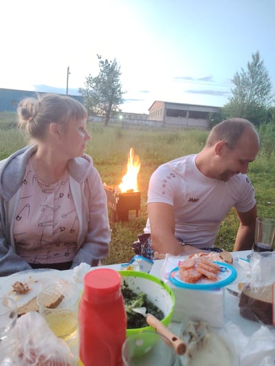 This media file depicts a casual outdoor gathering in a rural setting, possibly in Заямнаўскі сельскі Савет, Belarus. Two individuals, a woman with her hair in a bun and a bald man, are seated at a table laden with food. The woman, wearing a pink shirt with a safety pin pattern and a gray hooded jacket, looks to her right. The man, in a white t-shirt with "Jako" branding visible, is looking down and smiling. A portable grill with a fire is situated behind them, suggesting a barbecue or outdoor meal.
On the table, there are various food items, including sliced sausage on a blue and white plate, a bowl of green salad, bread, and glasses. A large red bottle, likely a ketchup or condiment dispenser, is prominently placed. The overall atmosphere appears relaxed and convivial, with the subjects engaged in enjoying a meal outdoors.
The scene is set in a grassy area with trees in the background, and what appear to be industrial or agricultural buildings further away. The lighting suggests it is either early morning or late evening, with the sky appearing a pale blue. The weather seems pleasant for an outdoor activity. The image captures a moment of leisure and social interaction.
