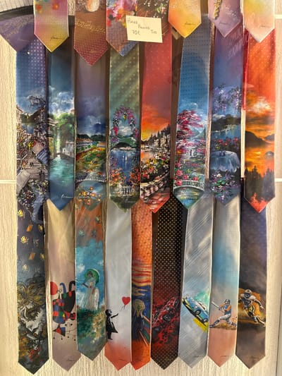 This media file displays a collection of hand-painted silk ties, likely for sale in a shop in San Giovanni, Italy, given the context. The ties are arranged horizontally, overlapping each other. Each tie features a unique, artistic design, suggesting a narrative or a theme related to art and culture.
The subjects vary greatly across the ties. Some depict serene landscapes with mountains and sunsets, others showcase vibrant floral gardens or picturesque Italian villages with architecture. There are also ties with more abstract or surreal imagery, including a figure flying a red balloon, a woman with an umbrella in a garden, and a stylized portrait of a face with elaborate hair. Some ties feature motifs related to music, such as musical notes or abstract representations of sound. A few ties depict sporting activities, like a baseball player or a football player. One tie displays a vintage car.
The overall mood is artistic and evocative, ranging from peaceful and romantic to playful and avant-garde. The time of day and weather are implied in some of the landscape scenes, with warm sunset hues in some and bright daylight in others. The location cues, such as the depiction of Italian villages, point to a European setting, consistent with the San Giovanni context.
Visible text includes "Hand Painted Ties" and "70€" on a small white tag attached to one of the ties. The artist's signature, "Giovanni," is visible on several of the ties, subtly incorporated into the designs.
