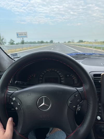 A first-person, eye-level perspective captures the interior of a Mercedes-Benz vehicle from the driver's seat, traveling on a multi-lane highway under a partly cloudy sky. The scene is identified as being in the vicinity of Malo Konare, Bulgaria, based on provided information and the presence of a billboard displaying "mobile.bg," a common Bulgarian website.
In the foreground, a black leather-wrapped steering wheel with prominent red stitching dominates the lower half of the frame. The steering wheel features a silver Mercedes-Benz logo at its center, with "SRS AIRBAG" embossed below it. Control buttons are present on both spokes: the left buttons include what appear to be menu navigation arrows and selection controls, while the right buttons feature volume adjustments (+/-) and phone call management (answer/end). A left hand, belonging to the driver, is partially visible gripping the lower-left portion of the steering wheel. The driver's leg, clad in blue denim fabric, is visible in the lower-right.
Behind the steering wheel, the instrument cluster is visible through the upper steering wheel gap. The speedometer needle indicates a speed between 80 and 90 km/h. The tachometer needle is positioned low, characteristic of cruising engine speed. A fuel gauge shows the tank approximately three-quarters full. A multi-function stalk, featuring "LIM" (limiter) and wiper symbols, protrudes from the left side of the steering column. A set of keys, including a metallic key fob, hangs from the ignition or a key slot near the right side of the steering column. Part of the vehicle's center console is visible on the far right, displaying buttons labeled "NAV," "CD," "SYS," and a rotary control knob. A black smartphone, held in a vertical mount, is partially visible on the extreme right edge of the frame. A blue fabric object or piece of material rests on the dashboard near the base of the windshield on the passenger side.
Outside the vehicle, a paved, multi-lane highway extends straight into the hazy distance, with white dashed and solid lane markings visible. A metal guardrail runs along the right shoulder of the highway. Sparse green vegetation, consisting of trees and bushes, lines both sides of the road. In the far distance on the left, a rectangular billboard displaying blurred text, discernibly "mobile.bg," is mounted on a pole. The upper portion of the image reveals a pale blue sky dotted with scattered white, cumulus-like clouds. The overall lighting suggests a clear daytime environment.