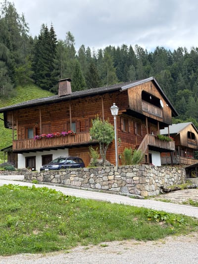 The image depicts a traditional wooden house in Sauris / Zahre, Italy. The house is situated on a hillside, with a dense forest of evergreen trees forming the backdrop. The building features multiple balconies adorned with flower boxes filled with colorful blossoms. A dark-colored station wagon is parked in front of the house, near a stone retaining wall. A paved path leads up to the property, and the surrounding area is covered in green grass and wildflowers. The sky is overcast, suggesting a cloudy day. The architecture of the house is characteristic of mountain dwellings, with its wooden construction and pitched roof. The scene conveys a sense of rural tranquility and alpine charm.