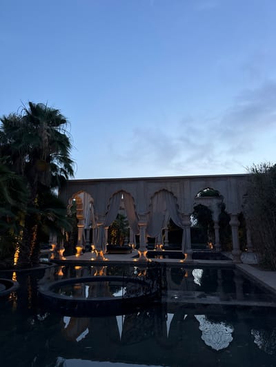 The image shows a courtyard in Ouled Jelal, Morocco, located at the coordinates 31.664, -7.869. It depicts a walled courtyard with a series of arches and fabric canopies. The arches are made of stone and feature intricately carved details. A body of water, presumably a pool, reflects the light from the sky and surrounding buildings. There are palm trees surrounding the courtyard, and the sky is a light blue, indicating twilight. The courtyard is empty of people, giving it a serene and tranquil atmosphere.