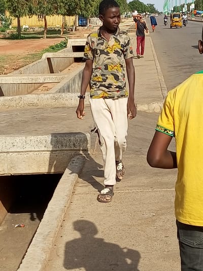 In Wuro Dole, Nigeria, a young man walks on a paved sidewalk beside a drainage ditch. He wears a camouflage t-shirt and light-colored pants, with sandals on his feet. To his right, another person in a yellow shirt is partially visible. In the background, other individuals and vehicles are present on a road, with trees and buildings lining the street. The scene appears to be a public area within the city.