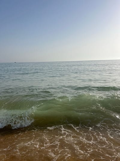 The image presents a forensic view of a beach scene in Almancil, Portugal, under clear skies. The sky occupies the upper third of the frame, displaying a light blue hue with subtle atmospheric haze at the horizon. The central and lower portions of the image are dominated by the ocean. The water exhibits a gradient, appearing lighter blue-green near the distant horizon and transitioning to a darker, more emerald green closer to the foreground. A single wave, positioned in the mid-foreground, is in the process of breaking, characterized by a visible curl of water and a crest of white foam. Beyond this wave, the water surface is relatively calm with minor ripples. The immediate foreground consists of damp, light brown sand with residual white foam patterns left by receding water. No individuals or human-made structures are present within the frame. A small, dark object, consistent with a boat, is faintly visible on the distant horizon in the upper left quadrant of the ocean.