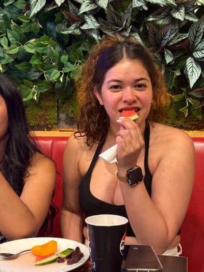 A young woman with curly brown hair is eating a slice of watermelon. She is wearing a black tank top and a watch on her left wrist. In front of her is a plate with fruit and a cup. She is seated at a table with a red upholstered bench. The background is a wall covered with lush green plants. The woman is in a restaurant. The location is Carretera De San Martín, Spain.