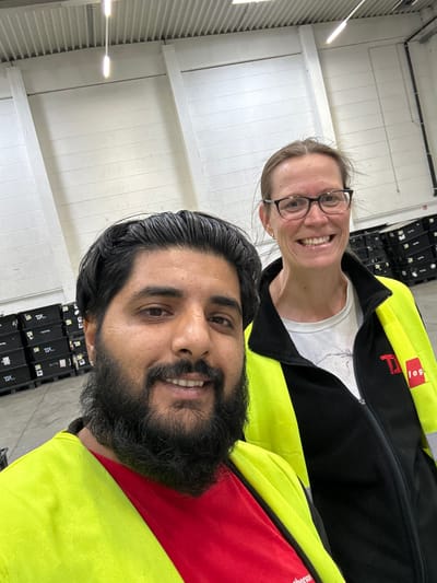 Two individuals, a man with dark hair and a beard, and a woman with blonde hair wearing glasses, are taking a selfie in what appears to be a warehouse environment. Both are wearing high-visibility safety vests over their clothing. The man is wearing a red shirt underneath his neon yellow vest. The woman is wearing a white t-shirt under her black jacket with neon yellow accents. Behind them, stacks of black boxes with the "TX LOGIS" logo are visible, suggesting a logistics or distribution center. The walls of the warehouse are white and ribbed, and fluorescent lights are visible on the ceiling. The scene appears to be indoors. The presence of the "TX LOGIS" branding strongly suggests the location is a facility operated by TX LOGIS. A search of TX LOGIS facilities indicates they have a presence in Schweitenkirchen, Germany, specifically at their distribution center at Flurstraße 1-3, 85304 Schweitenkirchen.