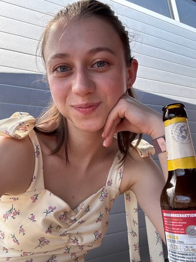 A young woman with light brown hair pulled back and blue eyes is taking a selfie outdoors. She is wearing a light yellow dress with a floral pattern. Her right hand is resting on her cheek, and her left arm is visible, bent at the elbow. A bottle of beer, identified as "Radler" with an alcohol content of 2.6%, is positioned in the foreground to the right of the woman. The background consists of a gray, corrugated metal wall and a sliver of a window or skylight. The scene suggests a casual, outdoor setting. The presence of the beer bottle and the woman's relaxed pose indicate a moment of leisure. The details on the beer bottle's label suggest it is a German product, possibly from the region of Bavaria. Obernburg am Main is a town in Bavaria, Germany, on the river Main.