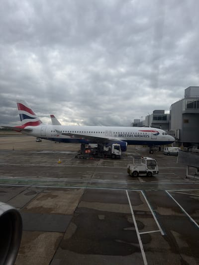 The image depicts a British Airways Airbus A320 series aircraft parked at an airport gate, likely at London Gatwick Airport, located in Crawley, United Kingdom. The aircraft is connected to ground service vehicles, including a refueling truck and a baggage towing tractor. The weather appears overcast with a cloudy sky. A portion of the airport terminal is visible adjacent to the aircraft's nose. Markings on the tarmac are visible including white taxiway lines. An engine nacelle is partially visible in the lower right corner of the image.