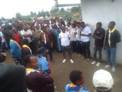 A large diverse group of people is gathered outside a building in a seemingly rural area. Some individuals wear what appear to be Scout-style neckerchiefs of varying colors. A few wear t-shirts with logos. Many are standing with their arms crossed. The landscape includes trees, a building, and a yellow vehicle visible in the distance, all under a cloudy sky.