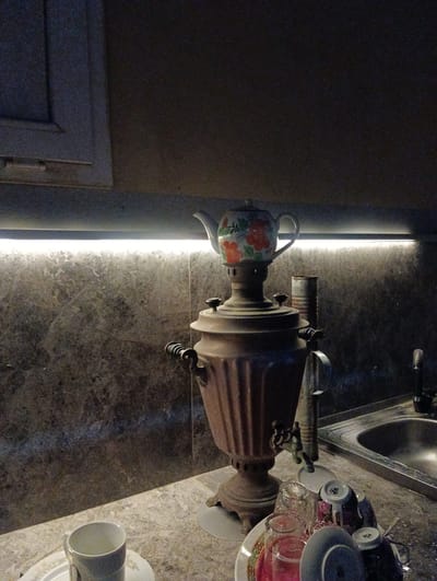 The image depicts an indoor scene with a traditional Russian samovar as the central object. The samovar is made of aged metal, likely brass or copper, showing signs of wear and patina. It sits on a marble-patterned countertop. A teapot with a floral design sits atop the samovar. To the right of the samovar is a sink with a faucet, also appearing to be made of metal. In the foreground, there are several teacups and saucers, some stacked, and a glass. The background features a marble-patterned wall with subtle lighting emanating from a strip light installed above the countertop. The overall setting suggests a domestic or hospitality environment, possibly a kitchen or a dining area. The location is identified as Türkan, Azerbaijan. There are no people visible in the image.