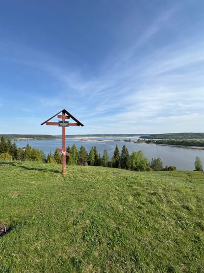 The image shows a wooden cross on a grassy hillside overlooking a body of water, presumably the Kama River, in Elkino, Russia. The cross is dark red in color with a triangular roof-like structure above the intersection of the vertical and horizontal beams. A plaque is affixed to the cross. The hillside is covered in green grass and some trees. The sky is blue with wispy clouds.