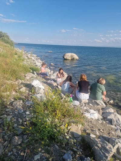 Four individuals are gathered on a rocky shoreline near a body of water. The scene appears to be a sunny day, with a clear blue sky and scattered white clouds. The water is a deep blue, with gentle ripples on its surface.
The individuals are positioned on the rocks, some facing the water and others looking towards each other. The person furthest back is wearing a green t-shirt and is sitting with their back to the camera. In front of them, another person is wearing a black t-shirt and white shorts, also seated. Closer to the water, two individuals are visible. One is wearing a light blue top and shorts, and the other, a pink top and shorts, is lying down. There are several objects scattered among the rocks, including a white plastic bag, a green bottle, and some white fabric.
The vegetation consists of dry grass and some green bushes with small yellow flowers growing amongst the rocks. The ground is covered with stones and pebbles of various sizes. The presence of rocks and the vast expanse of water suggest a natural, outdoor setting, possibly a lake or a large river. The overall mood seems relaxed and casual, with the individuals enjoying a day outdoors. The time of day is likely daytime, given the bright sunlight and clear sky. There is no visible text in the image.