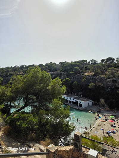 The image depicts a vibrant coastal scene in Llucmajor, Spain, likely Cala Pi given the distinctive white boathouse and surrounding cliffs. The turquoise water of the cove is dotted with swimmers and a yellow buoy, while a sandy beach hosts sunbathers and umbrellas. Dense green foliage and trees frame the cove, with a rocky, tree-lined cliff face rising behind it. In the foreground, a weathered stone post and railing suggest a viewing point overlooking the bay. The overall impression is of a popular summer destination on a bright, sunny day.