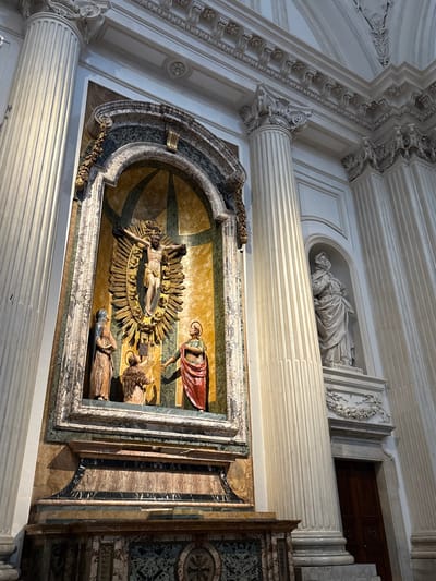 The image displays the richly decorated interior of the Cathedral of San Feliciano (Duomo di Foligno) in Foligno, Italy.
The central focus is an ornate, arched altarpiece set against a light-colored wall, flanked by two tall, fluted Corinthian columns of cream or white stone. The altarpiece features a deep niche containing a carved relief or sculpture of the Crucifixion. Jesus is depicted on the cross, made of a dark material, against a background of radiating golden sunburst rays. At the base of the cross, three figures are positioned: a standing female figure in a light brown robe on the left, a kneeling figure in a brown robe directly below the cross, and a standing male figure in a red tunic and light blue cloak with a raised hand on the right. The niche is framed by dark green, black, and white veined marble, with gold-colored leaf ornamentation along the arch. The background behind the figures within the niche is a lighter, patterned marble or fresco in yellow and green tones. Below the niche, a substantial altar structure made of multi-colored veined marble (brown, black, green) features a central circular medallion.
To the right of the main altarpiece, a smaller arched niche houses a white marble statue of a standing robed female figure, possibly a saint, on a rectangular pedestal. A faint inscription, possibly "B. ANGELINA VIVA," is visible on the pedestal. Below and slightly to the right of this statue, a dark wooden door with a rounded arch is partially visible.
Above the columns and altarpiece, an elaborate white entablature, adorned with classical moldings, rosettes, and dentils, extends horizontally. The ceiling, partially visible at the top-right, is vaulted and intricately decorated with white stucco relief work. The scene is illuminated by ambient light, creating subtle shadows that highlight the architectural details and sculptures. No living individuals are present in the image.
